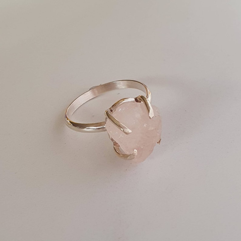 May include: A silver ring featuring a raw, light pink rose quartz gemstone. The stone is irregularly shaped and held in place by four prongs. The ring band is thin and polished, set against a white background.