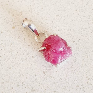 Peut inclure: Un pendentif en argent sterling avec une pierre précieuse de rubis brut. Le rubis est de couleur rose foncé et a une forme brute et naturelle. Le pendentif mesure environ 2,5 cm de long.