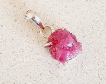 May include: A raw ruby gemstone pendant set in sterling silver. The ruby is a deep pink color and has a rough, natural shape. The pendant is approximately 1 inch in length.