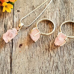 May include: A silver necklace with a pendant and two rings, all featuring rough-cut, light pink gemstones. The pendant hangs from a thin chain, while the rings have a simple band design. The jewelry is set against a rustic wooden background.
