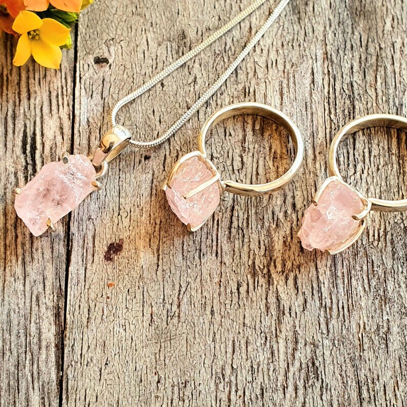 May include: A silver necklace with a pendant and two rings, all featuring rough-cut, light pink gemstones. The pendant hangs from a thin chain, while the rings have a simple band design. The jewelry is set against a rustic wooden background.