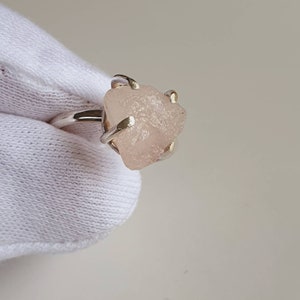 May include: A silver ring featuring a rough-cut, light pink gemstone. The stone is irregularly shaped and held in place by four prongs. The ring band is a simple, polished silver.