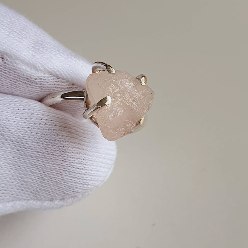 May include: A silver ring featuring a rough-cut, light pink gemstone. The stone is irregularly shaped and held in place by four prongs. The ring band is a simple, polished silver.