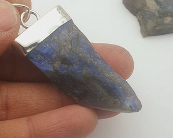 Puede incluir: Un colgante de plata con una piedra de labradorita azul oscuro en bruto. La piedra tiene una forma puntiaguda y tonos iridiscentes azules y verdes.