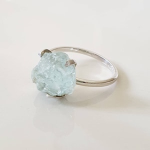 May include: A silver ring with a large, light blue aquamarine gemstone. The gemstone is raw and has a natural, irregular shape.
