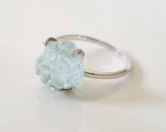 May include: A silver ring with a large, light blue aquamarine gemstone. The gemstone is raw and has a natural, irregular shape.