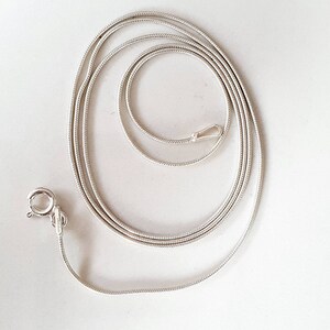 May include: A silver-colored snake chain necklace with a lobster clasp closure. The chain is coiled and lies on a white surface. The necklace is a delicate piece of jewelry.