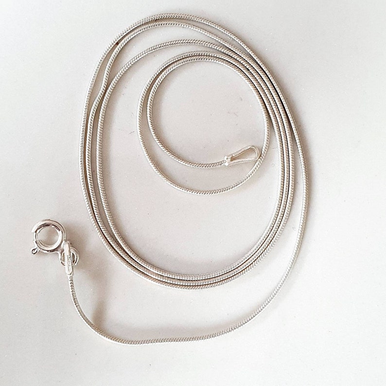 May include: A silver-colored snake chain necklace with a lobster clasp closure. The chain is coiled and lies on a white surface. The necklace is a delicate piece of jewelry.