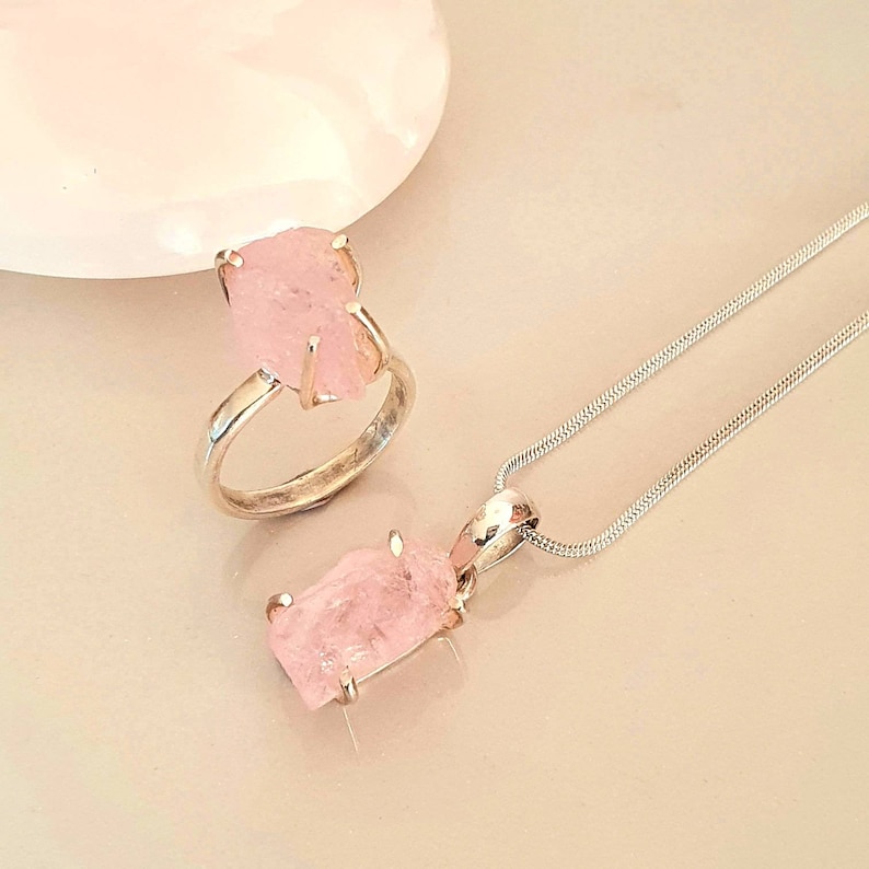 May include: A silver ring and pendant necklace set. The ring and pendant feature a rough-cut, light pink gemstone. The pendant hangs from a delicate silver chain. The jewelry is displayed on a light-colored surface.