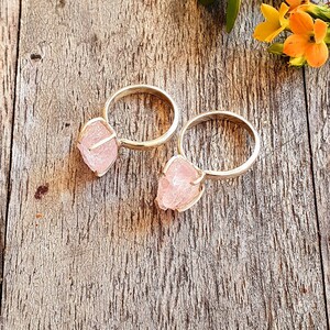 May include: Two silver rings with raw, light pink rose quartz stones. The rings are set on a rustic wooden surface, with a few orange and yellow flowers in the background. The rose quartz has a rough, natural texture.