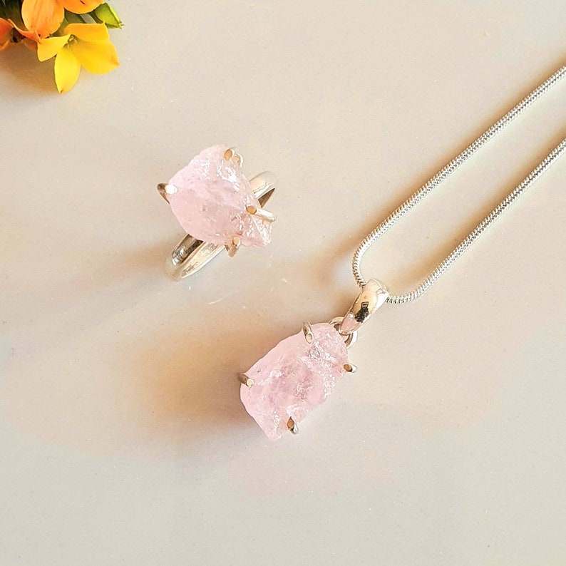 May include: A jewelry set featuring a ring and a pendant necklace. The ring and pendant both showcase a rough-cut, light pink gemstone held in place by silver-colored prongs. The necklace has a silver chain.