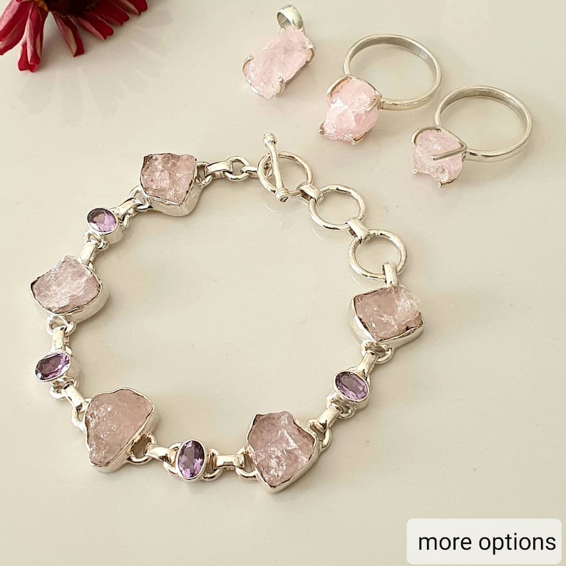 May include: A collection of jewelry featuring raw pink gemstones set in silver. The set includes a bracelet, two rings, and a pendant. The bracelet has large pink stones and smaller purple stones. The rings and pendant have similar pink stones.