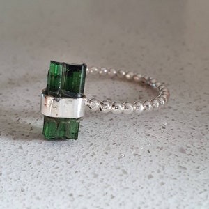 May include: A silver ring with a green tourmaline gemstone set in a bezel setting. The band is made of small silver beads.