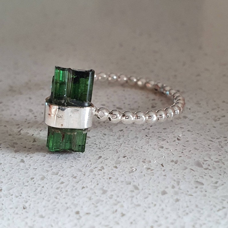 May include: A silver ring with a green tourmaline gemstone set in a bezel setting. The band is made of small silver beads.
