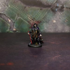 May include: A miniature figure of a character with dark hair, wearing a black and purple outfit, holding a red staff and a crossbow. The figure has intricate details, with metallic elements and a cape. The base is a dark, textured surface.