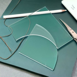 May include: Two clear glass pieces with rounded edges on a teal surface. The larger piece is rectangular and the smaller piece is a quarter circle.