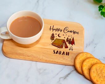 May include: A wooden serving tray with the words "Happy Camper" and a tent design. A white mug filled with a brown liquid sits on the tray, along with three biscuits. The name "SARAH" is also engraved on the tray.