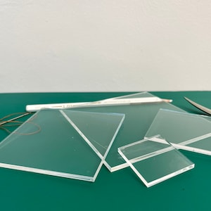 May include: Four clear acrylic squares of different sizes on a green surface. The largest square is partially overlapping a smaller square. A white pencil and a pair of scissors are visible in the background.