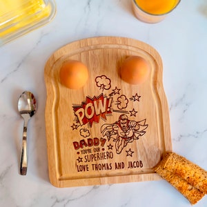 May include: A wooden breakfast board shaped like a slice of toast with two indentations for eggs. The board is engraved with a superhero graphic, the words "POW! Daddy You're Our Superhero Love Thomas and Jacob", and stars.
