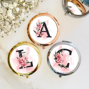 May include: Three round compact mirrors with a floral design and a letter on each. The mirrors are gold, silver, and rose gold. The letters are A, L, and C.