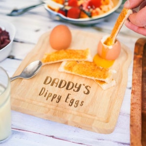 May include: A wooden serving board with the text "Daddy's Dippy Eggs" engraved on it. There are two soft boiled eggs, one with the top peeled off, and several slices of toast on the board.