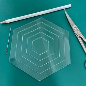 May include: A clear plastic hexagon template with four concentric hexagons. The template is on a green surface with a white pencil, a needle, and silver scissors.