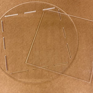 May include: Two clear acrylic sheets, one circular and one square, with cut-out lines for scoring. The sheets are lying on a brown surface.