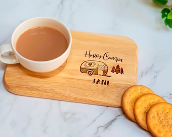 May include: A wooden serving board with a cup of tea and biscuits. The board is engraved with a camper design, the words "Happy Camper", and the name "JANE". The cup is white, and the tea is light brown. Three biscuits are on the right.