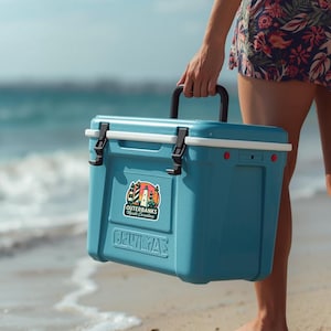 Puede incluir: Una nevera r&iacute;gida de color azul claro con un asa superior negra y cierres negros. Una pegatina en la parte frontal muestra tres faros y el texto Outerbanks North Carolina. La nevera tiene un borde blanco y un logotipo moldeado en el frente.