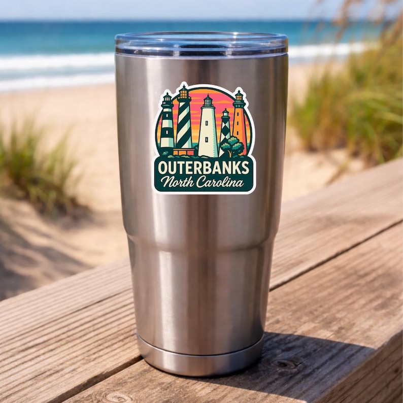 Puede incluir: Un vaso de acero inoxidable con una pegatina colorida de cinco faros contra un cielo al atardecer. La pegatina incluye el texto OUTERBANKS North Carolina en letras blancas sobre un fondo verde azulado oscuro, con una tapa de pl&aacute;stico transparente.