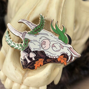 Op de afbeelding: Gedetailleerde emaille pin met een hertenschedel ontwerp, geweien, laurierbladeren en herfstthema-elementen. De pin heeft een gouden omtrek en is geplaatst tegen een crèmekleurig schedelmasker.