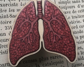 May include: A wooden pin in the shape of lungs, featuring a detailed anatomical illustration in shades of red and brown. The pin is set against a backdrop of vintage newspaper text, creating a unique, artistic aesthetic.