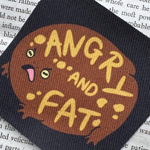 May include: A square black fabric patch featuring a brown, round toad illustration with yellow spots, yellow eyes, and a pink tongue. The text ANGRY AND FAT is printed in yellow across the centre of the toad's body in a bold, hand-drawn style font.