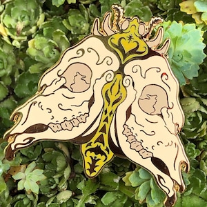 May include: Gold-tone metal pin featuring a butterfly with two skull heads. The butterfly has a yellow and green design with intricate details. The pin is set against a background of green foliage.