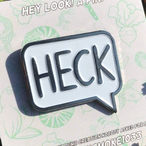 May include: A white enamel pin with a black speech bubble that says "HECK" on it. The pin is on a white card with a green and white floral design. The card also has the text "HEY LOOK! A PIN!" and "ANOTHER DUMB CREATION NOBODY ASKED FOR BY @BLACKSMOKE1033".