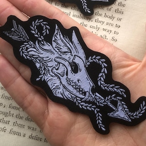 Może przedstawiać: Dwie czarne, haftowane naszywki z czaszką, strzałą i elementami kwiatowymi. Naszywki są w kolorze jasnoszarym i białym, z precyzyjnymi detalami. Naszywki są trzymane w dłoni.