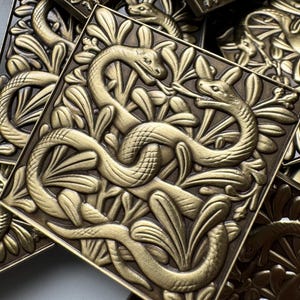May include: A collection of square, metallic gold-toned tiles featuring a raised, embossed relief design of two intertwined snakes surrounded by stylised foliage. The tiles have a dark, recessed background that highlights the detailed snake motifs.