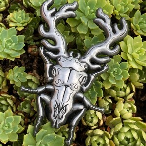May include: A silver metal pendant in the shape of a stylized beetle with large antlers. The pendant features detailed patterns and is set against a backdrop of green succulents.