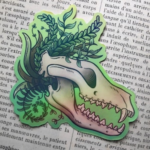 May include: A holographic sticker featuring a stylized animal skull with green and pink hues, adorned with green foliage. The sticker is placed on a vintage book page.