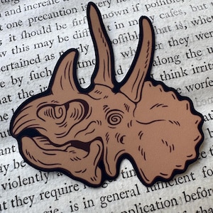 May include: A tan-colored sticker featuring a stylized illustration of a triceratops skull with dark brown outlines and detailed line work, showcasing two prominent brow horns and a large frill, set against a background of vintage text.