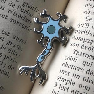Può includere: Spilla a forma di neurone color argento con design in smalto blu chiaro e blu scuro. La spilla è appoggiata sulle pagine di un libro aperto. Un accessorio a tema scientifico.