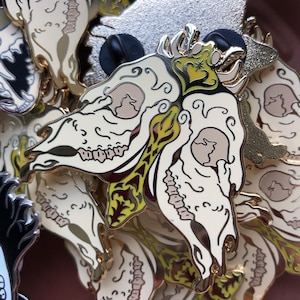 May include: A close-up of several enamel pins featuring a stylized skull design with intricate details. The skulls are painted in a cream color with yellow and green accents. The pins are arranged in a pile, with the back of one pin visible, showing a gold-colored metal backing.