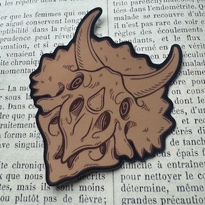 May include: A tan-colored sticker featuring a detailed illustration of a triceratops skull with dark brown outlines and shading. The dinosaur skull is depicted in profile with its characteristic three horns and a large, bony frill behind the head.