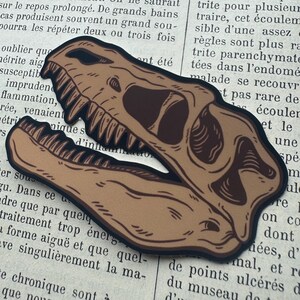 May include: A tan and dark brown sticker featuring a stylized illustration of a dinosaur skull with an open jaw, showing sharp teeth and deep eye sockets, outlined in a thick black border against a background of printed French text.
