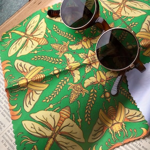 Może przedstawiać: Okrągłe okulary przeciwsłoneczne z brązowymi oprawkami spoczywają na zielonej tkaninie z żółtymi owadami i kwiatowymi wzorami. Okulary mają ciemne soczewki. Tkanina leży na otwartej książce z francuskim tekstem.