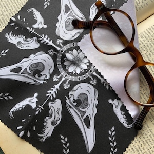 Puede incluir: Un paño de limpieza negro con un patrón repetido de calaveras de pájaros blancas y elementos botánicos. Gafas marrones descansan sobre el paño, que está sobre un libro abierto.