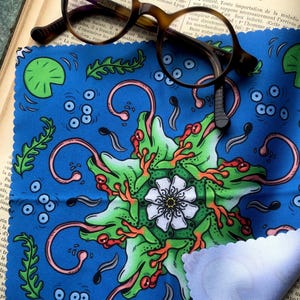 Puede incluir: Gafas redondas marrones descansan sobre un paño azul con un diseño caprichoso de ranas y nenúfares. Se ve una esquina de un paño de limpieza blanco. El fondo incluye un libro abierto con texto en francés.