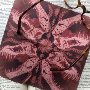 May include: A maroon and black patterned microfiber cloth featuring a repeating design of dinosaur skulls and floral elements. Tortoiseshell spectacles rest on the cloth, which is placed on an open book.