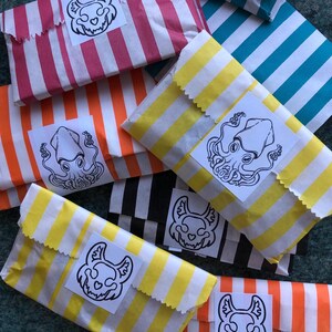 Könnte beinhalten: Eine Sammlung gestreifter Papiertüten in verschiedenen Farben, darunter Rosa, Orange, Gelb, Türkis und Schwarz. Jede Tüte ist versiegelt und mit einem weißen Aufkleber mit einer schwarz-weißen Illustration versehen. Die Tüten sind zusammengefasst.