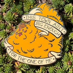 May include: A yellow enamel pin featuring a cartoon character in a cowboy hat. The pin includes a banner with the text "THIS TOWN AIN'T BIG ENOUGH FOR THE ONE OF ME". The pin is displayed on a bed of green plants.